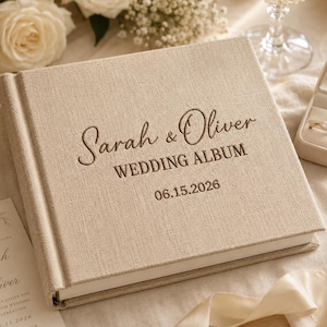 Puede incluir: Un álbum de fotos cuadrado con cubierta de lino beige que presenta el texto en relieve Sarah & Oliver WEDDING ALBUM 06.15.2026. El álbum tiene un acabado de tela texturizada y un lomo resistente, ofreciendo una apariencia limpia y neutra.