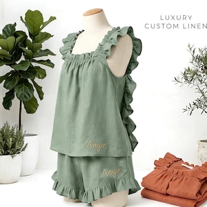 Puede incluir: Conjunto de pijama de lino verde salvia con volantes, exhibido en un maniquí. El conjunto incluye una camiseta sin mangas y pantalones cortos, ambos bordados con el nombre "Angie". El texto "LUXURY CUSTOM LINEN" es visible. También se ve una pila de pijamas naranjas.
