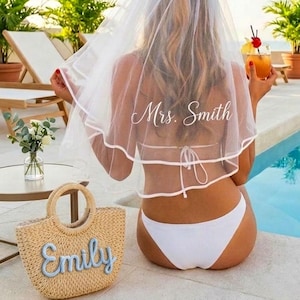 Pode incluir: Uma noiva usando um véu branco com "Mrs. Smith" impresso, sentada à beira de uma piscina. Ela está usando um biquíni branco e segurando um coquetel. Uma bolsa de praia de palha com "Emily" bordado está por perto.