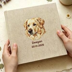 Op de afbeelding: Een vierkant, beige fotoalbum met een textuur van stof, voorzien van een gedetailleerde illustratie van een golden retriever-gezicht op de voorkant. Onder de illustratie van de hond staat de tekst Deegan en de jaren 2019-2026 in bruin.