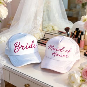 Puede incluir: Dos gorras de camionero blancas, una con "Bride" y otra con "Maid of Honor" en escritura rosa. Tienen la parte trasera de malla y una visera curva. Las gorras están sobre una superficie blanca, con maquillaje y flores en el fondo.