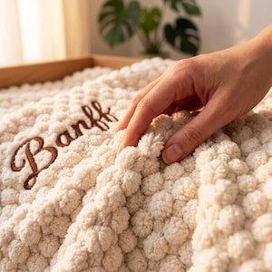 以下が含まれることがあります： 「Banff」という文字が茶色の筆記体で刺繍された、クリーム色のテクスチャーのあるボブルニットブランケット。生地全体にポンポンのような隆起した表面の質感があり、柔らかく触り心地の良い外観を演出しています。