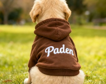 Może przedstawiać: Szczeniak golden retriever w brązowej bluzie z kapturem z napisem "Paden" wyhaftowanym na plecach białą czcionką. Pies siedzi na zielonej trawie, tyłem do aparatu.