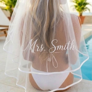 May include: A white bridal veil with a double-layered design and a satin trim. The veil is embroidered with the name "Mrs. Smith" in elegant script. The bride is wearing a white bikini.