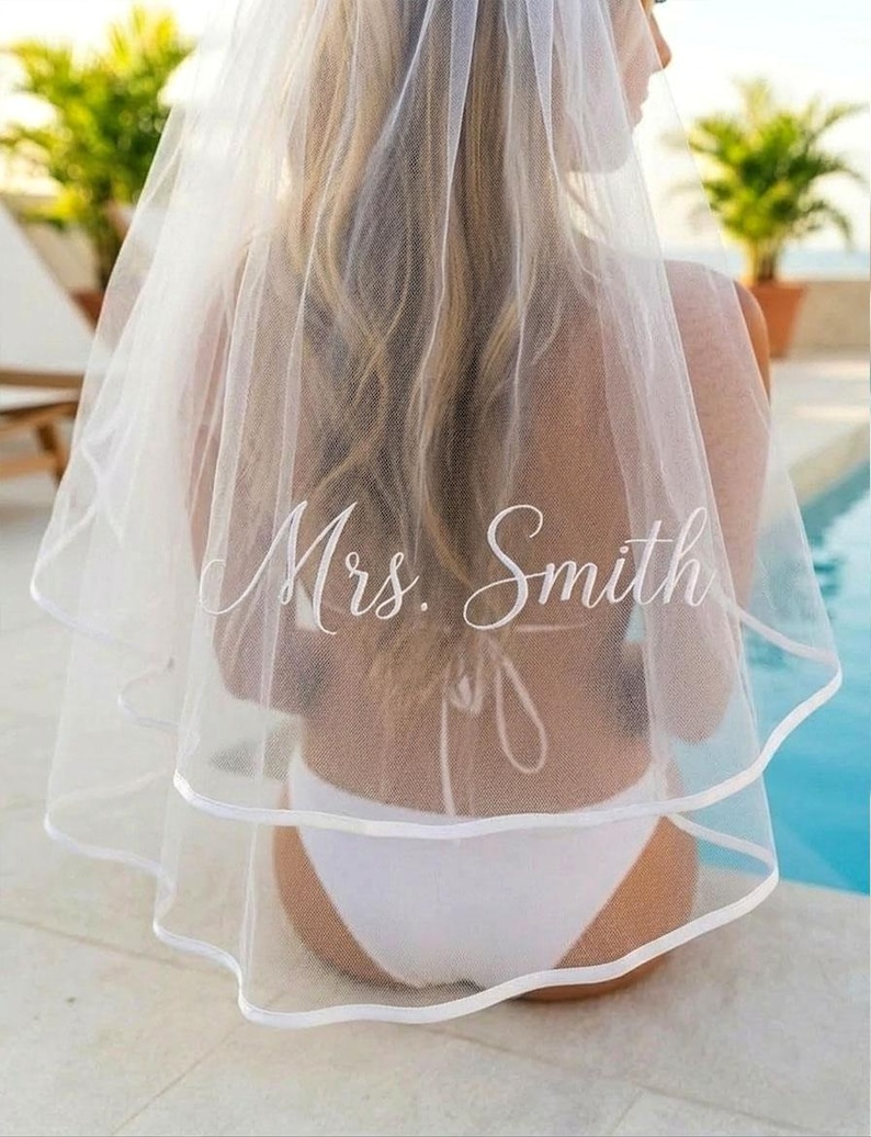 May include: A white bridal veil with a double-layered design and a satin trim. The veil is embroidered with the name "Mrs. Smith" in elegant script. The bride is wearing a white bikini.