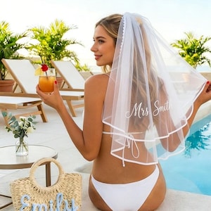 May include: A white bridal veil with the text "Mrs Smith" in script. A straw tote bag with the name "Emily" in blue script. The image shows a woman wearing a white bikini and holding a cocktail near a pool.