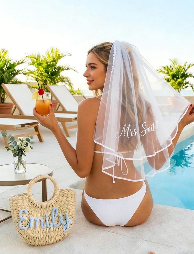May include: A white bridal veil with the text "Mrs Smith" in script. A straw tote bag with the name "Emily" in blue script. The image shows a woman wearing a white bikini and holding a cocktail near a pool.
