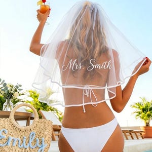 May include: A bride-to-be wearing a white veil with "Mrs. Smith" printed on it, and a white bikini. She is holding a cocktail with a cherry garnish. A straw tote bag with the name "Emily" is in the foreground.