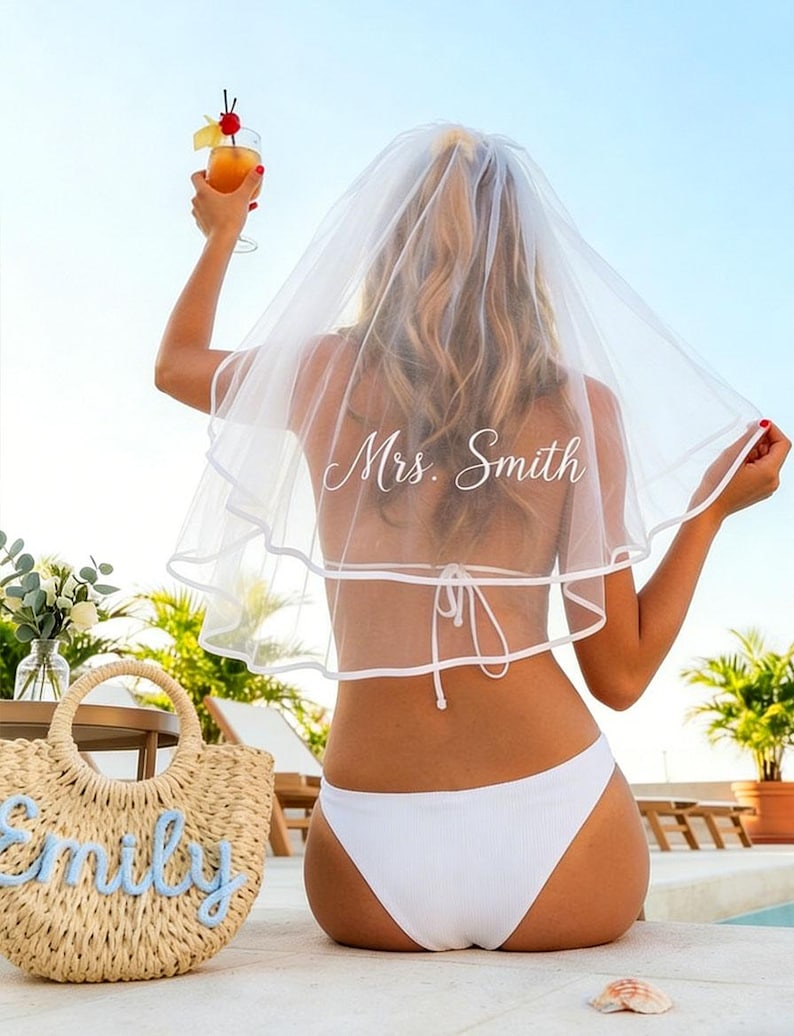 May include: A bride-to-be wearing a white veil with "Mrs. Smith" printed on it, and a white bikini. She is holding a cocktail with a cherry garnish. A straw tote bag with the name "Emily" is in the foreground.