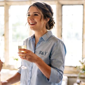 May include: A pale blue button-down shirt with the embroidered letters "B&Y" in dark teal. The shirt is worn by a person holding a champagne flute. The image is taken indoors with natural light.