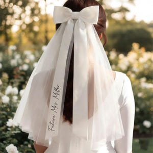 May include: A white bridal veil with a large bow at the top. The veil has a sheer, flowing design and the text "Future Mrs. Miller" is printed on the side. The background is a soft focus of greenery and white flowers.