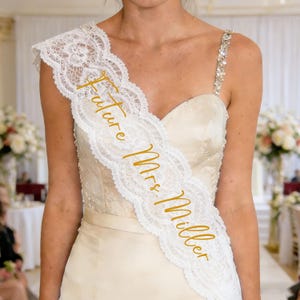 May include: A white lace sash with the text "Future Mrs. Miller" in gold script. The sash is draped over a cream-coloured wedding dress. The dress has a jewelled strap and a sweetheart neckline. The sash is a bridal accessory.