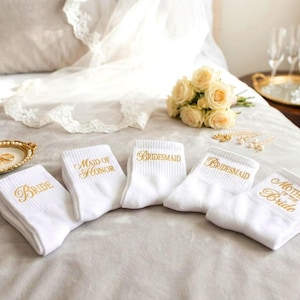 Puede incluir: Calcetines blancos bordados con texto dorado, incluyendo "Novia", "Dama de honor", "Bridesmaid" y "Madre de la novia". Los calcetines están colocados sobre una cama con anillos de boda, flores y copas de champán.