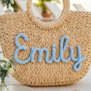 May include: A woven straw tote bag with a circular handle. The name "Emily" is spelled out in light blue rope across the front of the bag. The bag is beige and appears to be sitting on a white surface.
