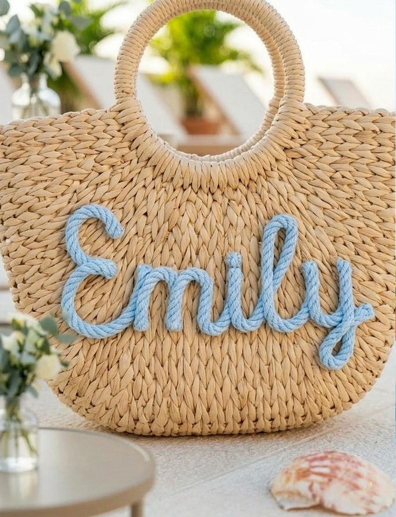 May include: A woven straw tote bag with a circular handle. The name "Emily" is spelled out in light blue rope across the front of the bag. The bag is beige and appears to be sitting on a white surface.