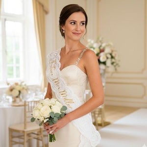 Puede incluir: Una futura novia lleva una banda de encaje blanco con texto dorado que dice "Future Mrs. Miller". Sostiene un ramo de rosas blancas y vegetación. La novia lleva un vestido de novia blanco con tirantes adornados.