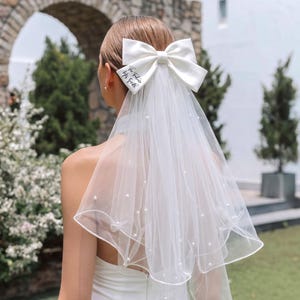 May include: A white bridal veil featuring a large satin bow at the top with the text The Future Mrs. Smith printed in black script. The sheer tulle fabric is adorned with scattered white pearls and has a finished edge, creating a layered look.