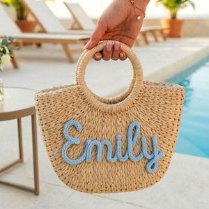 May include: A woven straw tote bag with a circular handle is held up by a person. The bag features the name "Emily" in light blue script across the front. The background includes a pool and lounge chairs.