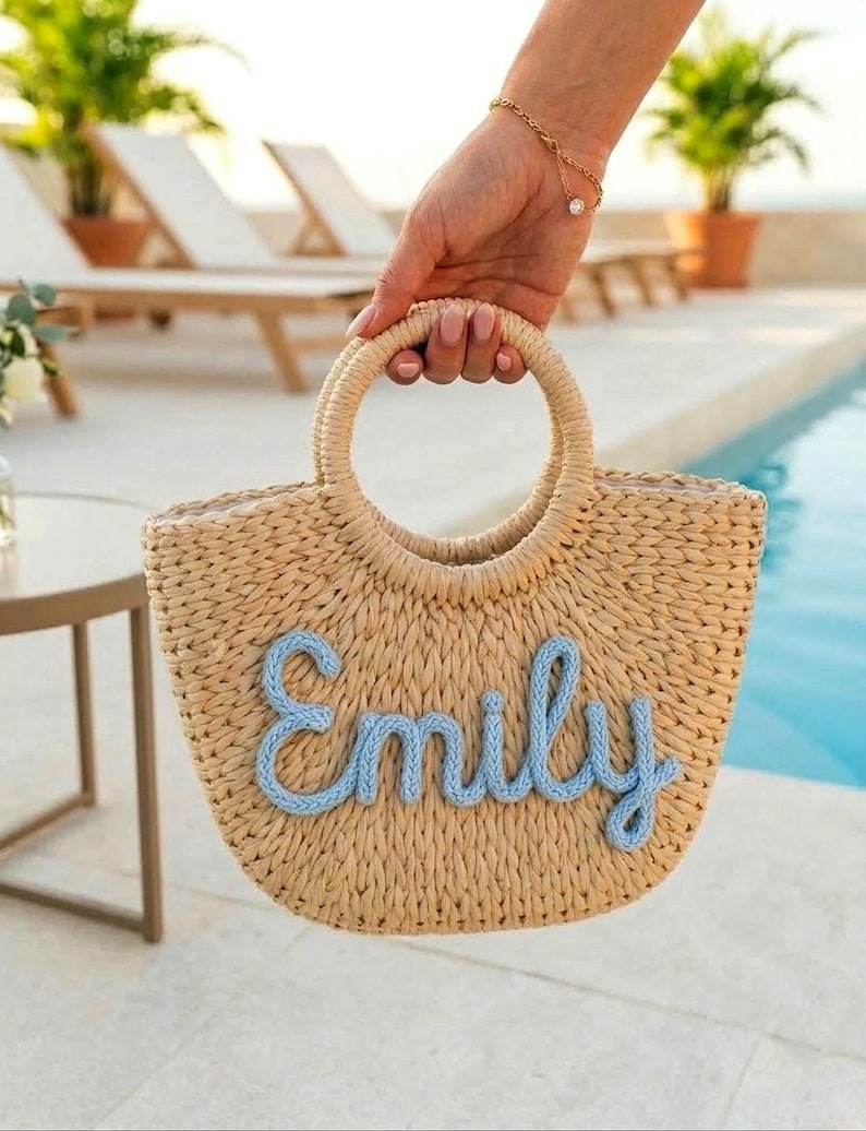 May include: A woven straw tote bag with a circular handle is held up by a person. The bag features the name "Emily" in light blue script across the front. The background includes a pool and lounge chairs.