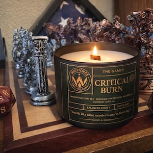 May include: A lit black candle labeled "Critical Burn" with the text "The Gamer" on a wooden surface. The candle is surrounded by chess pieces, dice, and other game-related figurines. The candle's scent is described as mountain coffee, midnight, and smoked vanilla.
