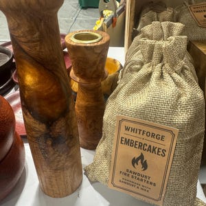 Könnte beinhalten: Kerzenhalter aus Holz und ein Jutesack mit der Aufschrift "Whitforge Embercakes" mit einer Flammen-Grafik. Der Beutel enthält Sägemehl-Anzünder, handgefertigt mit Sägemehl und Wachs. Die Kerzenhalter sind aus Holz mit natürlichem Finish.