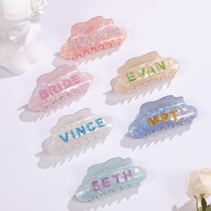 Op de afbeelding: Een verzameling iriserende haarclips in verschillende kleuren, elk met een naam of woord in glitter. De clips zijn in roze, blauwe en witte tinten, met namen als "BRIDE" en "SETH".