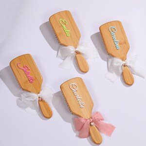 May include: Four personalized wooden hairbrushes with names in various colors. Each brush has a wooden handle and is adorned with a ribbon bow. The names are "Emilia", "Carolina", "Juliette", and "Charlotte".