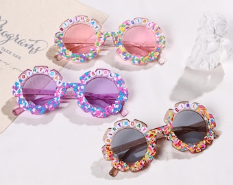 May include: Four pairs of colorful sunglasses with flower-shaped frames. The frames are decorated with sprinkles and the words "BIRTHDAY GIRL". The lenses are pink, purple, and gray. A fun accessory for a birthday celebration.