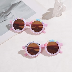 May include: Two pairs of pink sunglasses with brown lenses. One pair has the name "CHARLENE" spelled out in letter beads, the other "KINGSTON". Both pairs have pearl and heart-shaped embellishments.