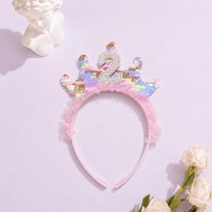 Könnte beinhalten: Ein rosa Stirnband mit einer paillettenbesetzten Krone mit der silbernen Glitzerzahl "2". Die Krone hat pastellfarbene Pailletten und goldfarbene Akzente. Ein rosa Tüllbesatz verleiht dem Stirnband eine weiche Note, ideal für einen Geburtstag.
