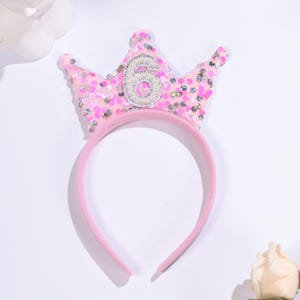 May include: A pink headband with a sequined crown featuring the number 6 in silver. The crown is decorated with pink sequins and has a soft, rounded shape. The headband is a solid, pale pink color.