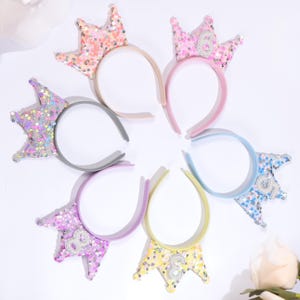 May include: A collection of colorful sequined crown headbands. Each headband features a different color crown, including pink, peach, purple, blue, and yellow. Some crowns have numbers. The headbands are designed for children's parties or special occasions.