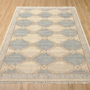 Op de afbeelding: Rechthoekig vloerkleed met een geometrisch ruitpatroon in lichtblauw en beige. Het kleed heeft een rand met een bloemenpatroon en franjes langs de randen. Het kleed is gemaakt van geweven materiaal.