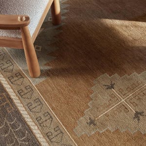 Peut inclure: Une section d'un tapis à motifs dans les tons de marron, beige et gris. Le tapis présente des motifs géométriques et des bordures. Un pied de chaise en bois et une partie de l'assise sont visibles dans le coin supérieur gauche.