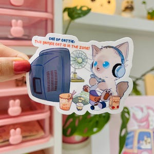 May include: A sticker featuring a cartoon cat with blue eyes wearing headphones, playing a video game. The sticker includes the text "CALL OF CATTIE: This GAMER CAT IS IN THE ZONE!" and images of snacks and a retro TV.