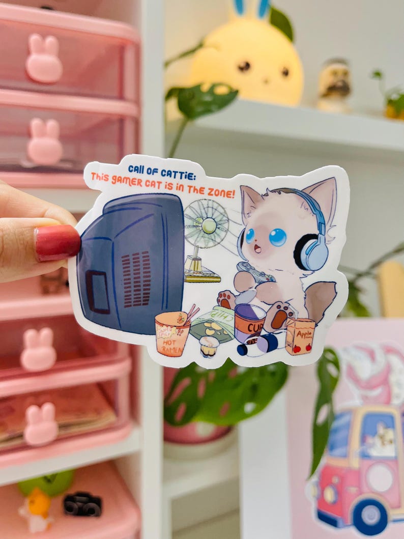 May include: A sticker featuring a cartoon cat with blue eyes wearing headphones, playing a video game. The sticker includes the text "CALL OF CATTIE: This GAMER CAT IS IN THE ZONE!" and images of snacks and a retro TV.
