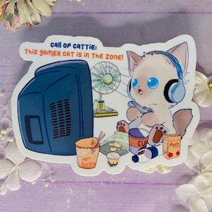 May include: A sticker featuring a cartoon cat with blue eyes wearing headphones and playing a video game. The text reads "CALL OF CATTIE: This GAMER CAT is in THE ZONE!" with images of ramen, chips, and an apple juice box.