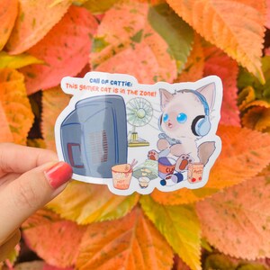May include: A sticker featuring a cartoon cat with blue eyes wearing headphones and playing a video game. The sticker has the text "CALL OF CATTIE: This gamer CAT is in the ZONE!" and includes images of a computer, fan, and snacks.