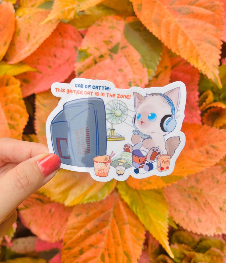 May include: A sticker featuring a cartoon cat with blue eyes wearing headphones and playing a video game. The sticker has the text "CALL OF CATTIE: This gamer CAT is in the ZONE!" and includes images of a computer, fan, and snacks.