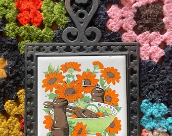 May include: A vintage trivet with a decorative metal frame and a ceramic tile insert. The tile features a floral design with orange sunflowers, a green bowl, and brown salt and pepper shakers. The trivet is set against a colourful crocheted background.