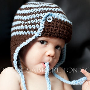 May include: A baby wearing a brown and blue crocheted hat with a blue button. The hat has a long strap that the baby is holding in their hand.