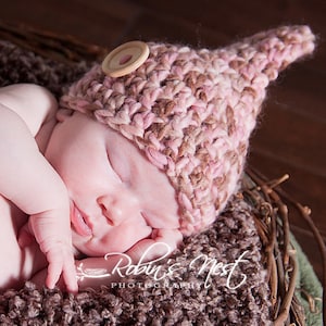Peut inclure: Un nouveau-né portant un bonnet en crochet rose et marron avec un bouton en bois sur le dessus. Le bébé dort paisiblement dans un nid brun.