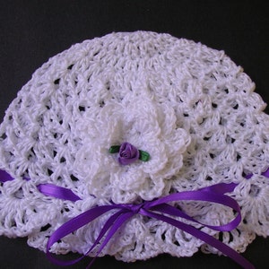 May include: White crocheted baby hat with a purple satin ribbon and a white crocheted flower with a purple center.