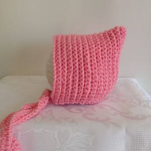 May include: A pink crocheted baby bonnet with a long tie. The bonnet is made with a simple stitch pattern and has a pointed crown.