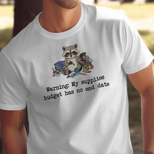 May include: A white short-sleeve t-shirt featuring a graphic of a raccoon wearing glasses surrounded by craft supplies. Below the graphic, the text reads: Warning: My supplies budget has no end date. The design includes yarn, scissors, and paint.
