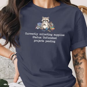 May include: A navy blue short-sleeve t-shirt featuring a graphic of a raccoon surrounded by craft supplies and the white text: Currently collecting supplies Status: Unfinished projects pending. The shirt has a crew neckline and rolled sleeves.