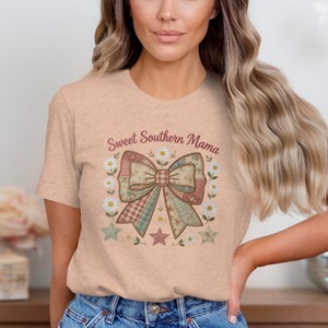 以下が含まれることがあります： 「Sweet Southern Mama」の文字とパッチワークのリボンデザインがプリントされた、ピーチ色のTシャツ。リボンの周りには、デイジー、星、花のボーダーがあしらわれています。柔らかい素材で作られたシャツです。