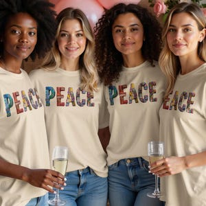 May include: Four people wearing cream-colored t-shirts with the word "PEACE" in colorful, patterned letters. They are holding champagne flutes and smiling. The background includes pink balloons and flowers.