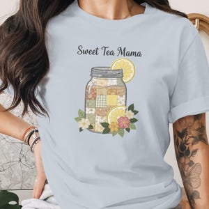 May include: Light blue t-shirt with the text "Sweet Tea Mama" above a mason jar illustration. The jar contains a patchwork design and is garnished with lemon slices and flowers. The shirt is a soft, comfortable material.