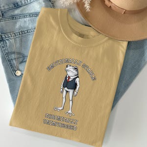 Könnte beinhalten: Senfgelbes T-Shirt mit einer Cartoon-Fröschin, die einen Anzug und eine Krawatte trägt. Der Text "EMOTIONALLY STABLE" wölbt sich über dem Frosch, darunter steht "CHRONICALLY UNIMPRESSED". Das Shirt liegt auf einer blauen Jeans und einem beigen Hut.